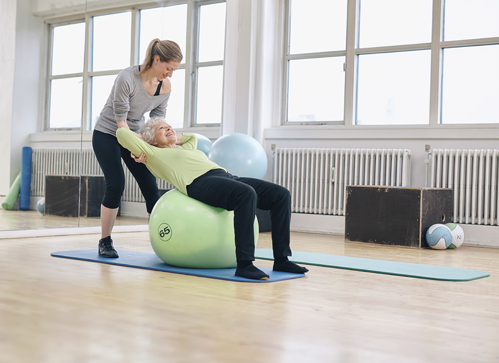 Female coach helping senior woman exercising in gym. Elder woman working out on pilates ball at health club being assisted by her personal trainer.